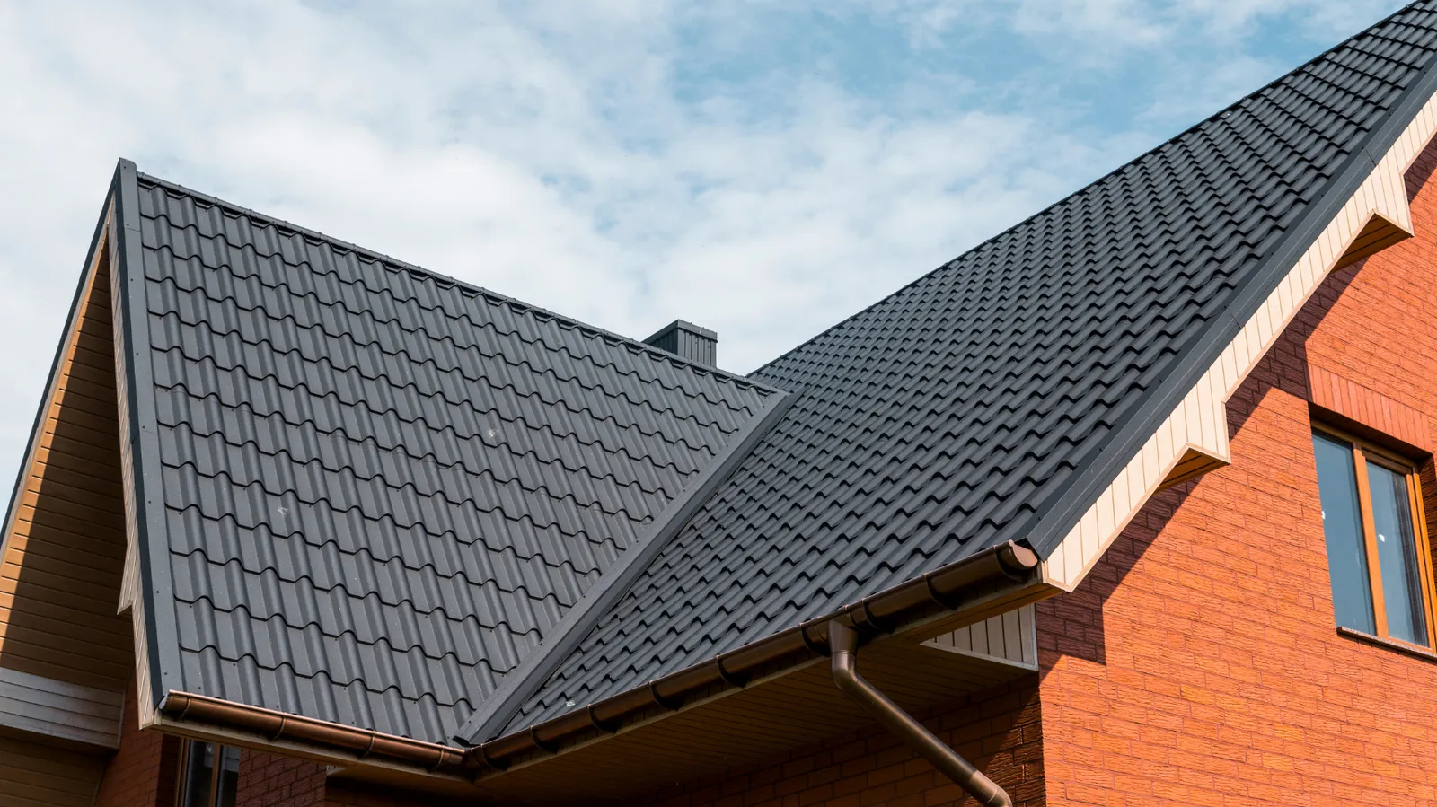 Dark metal tile roofing on a brick house under a partly cloudy sky